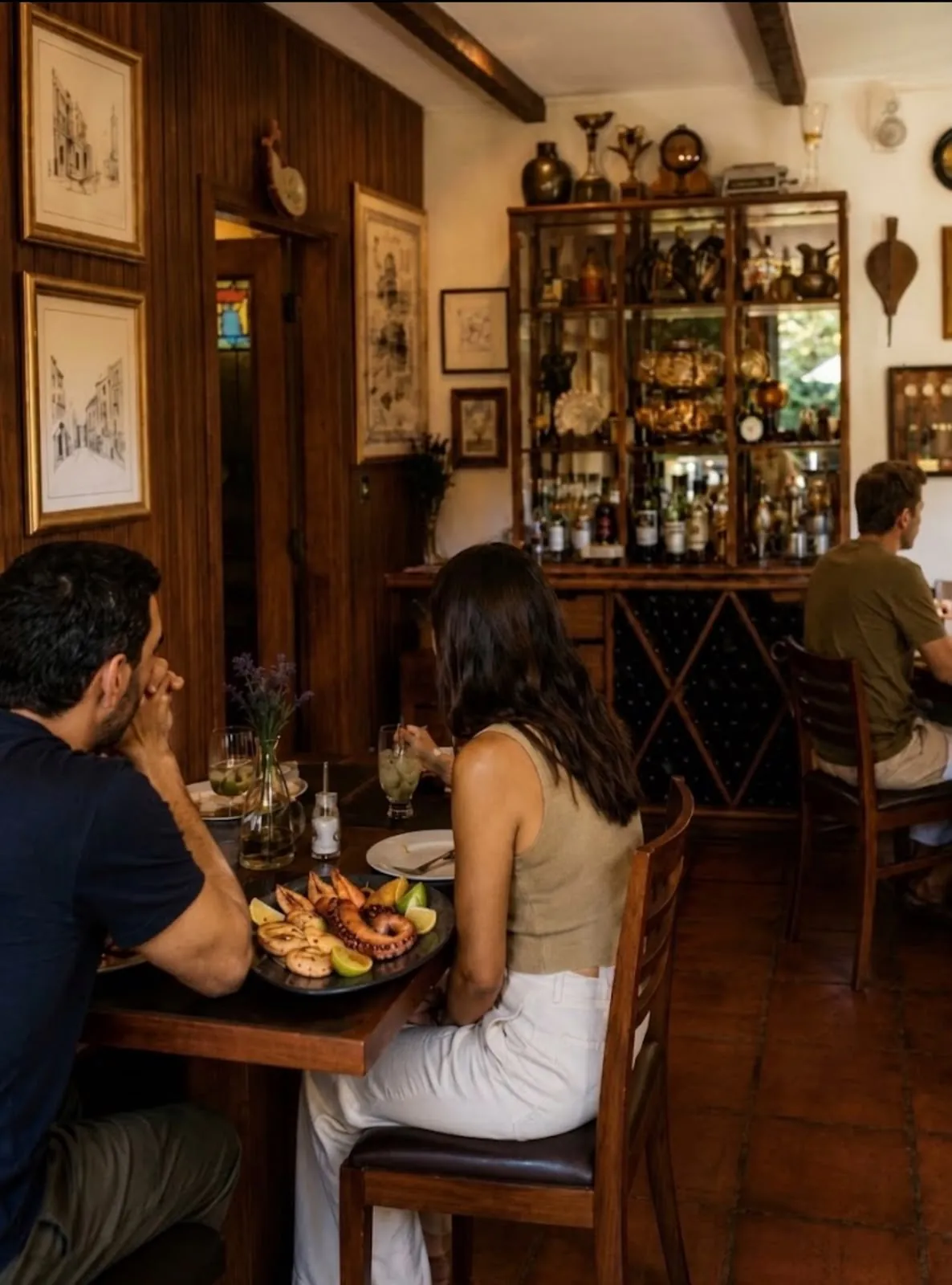 Casal almoçando no salão do Empório Araçá, com um prato de polvo à mesa e a cristaleira de bebidas ao fundo