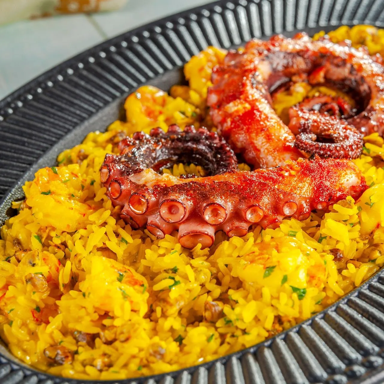 Tentáculos de polvo grelhados sobre arroz amarelo com frutos do mar, servidos em travessa escura